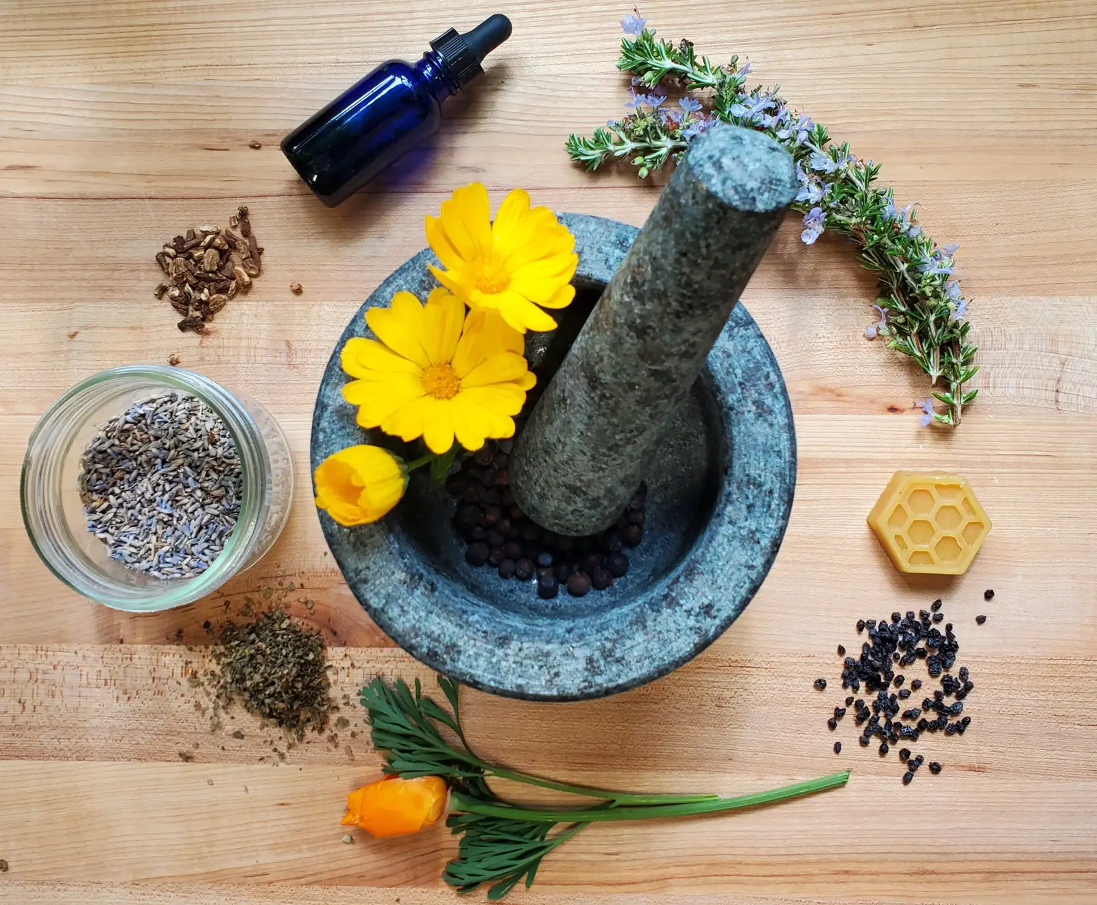 Mortar and pestle with calendula, lavender, rosemary, and herbal ingredients on a wooden surface